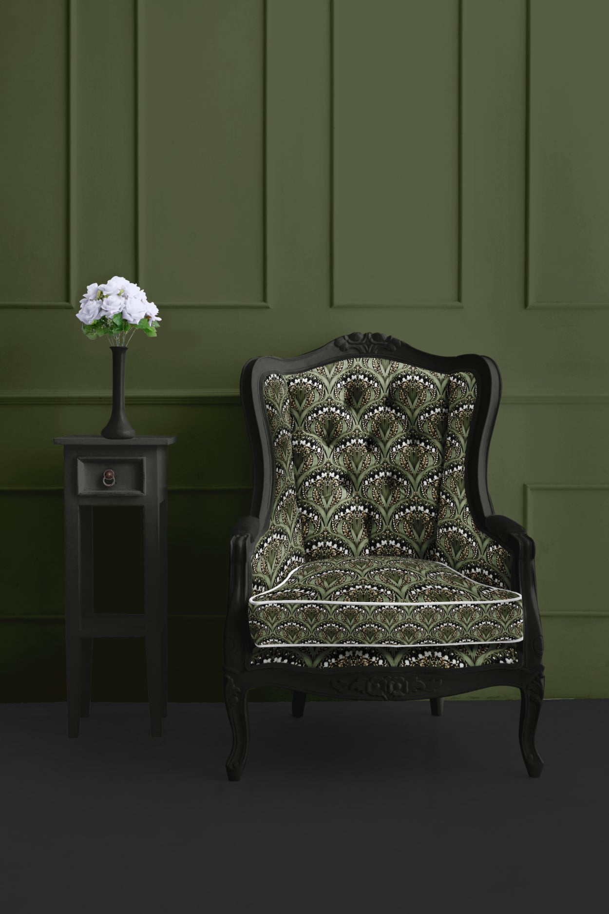 Dark green interior with an ornate black armchair upholstered in a green Art Nouveau floral pattern, placed beside a black side table with a tall vase of white flowers against a paneled green wall.