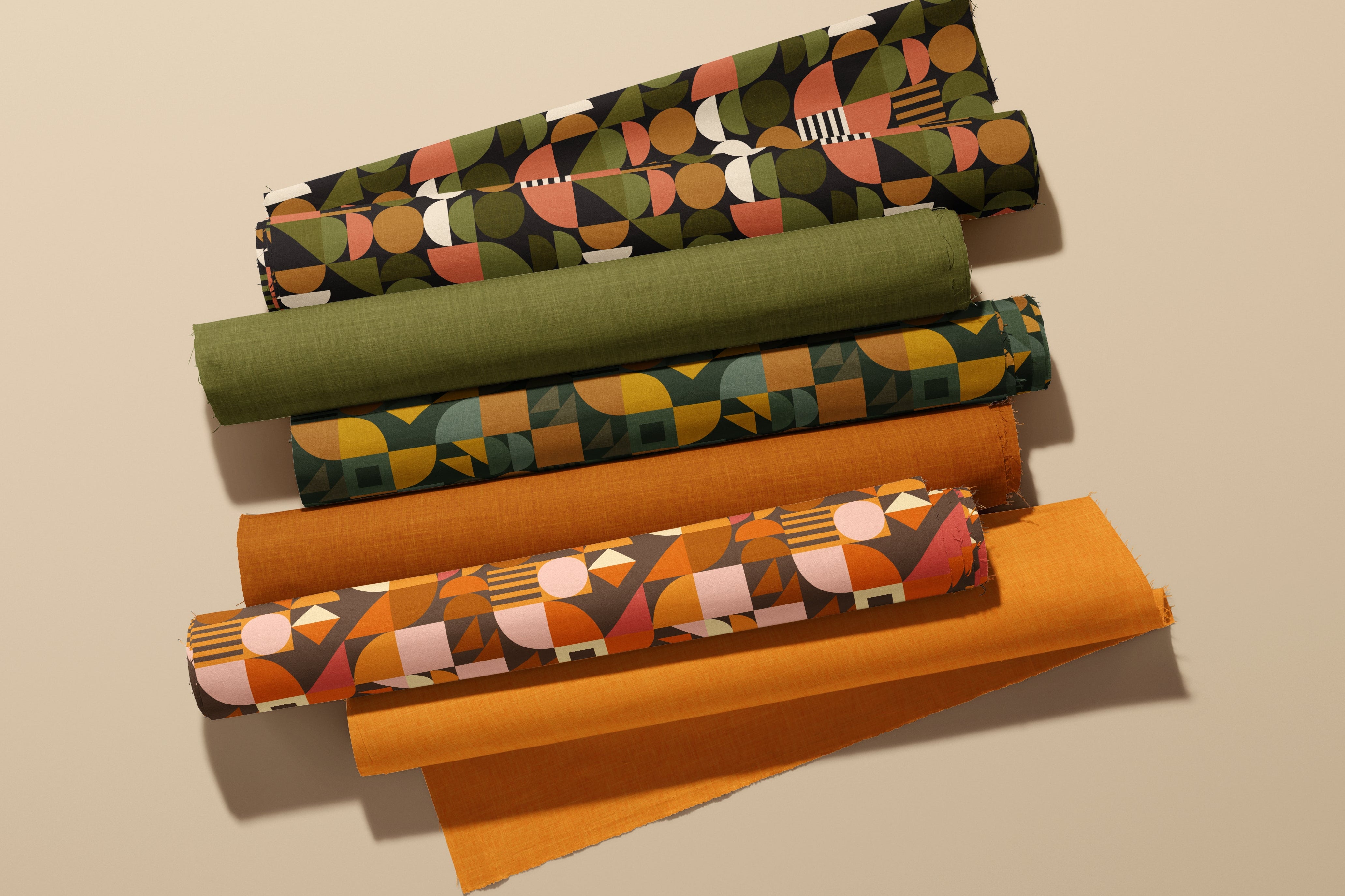 six rolls of patterned and solid fabric in earthy tones, including olive green, mustard yellow, terracotta orange,  geometric designs, arranged diagonally on a warm beige background.