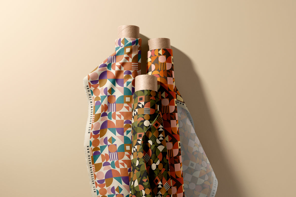 Three rolls of patterned fabric with kraft paper ends, featuring geometric designs in orange, purple, green, and blue, arranged against a solid beige background.