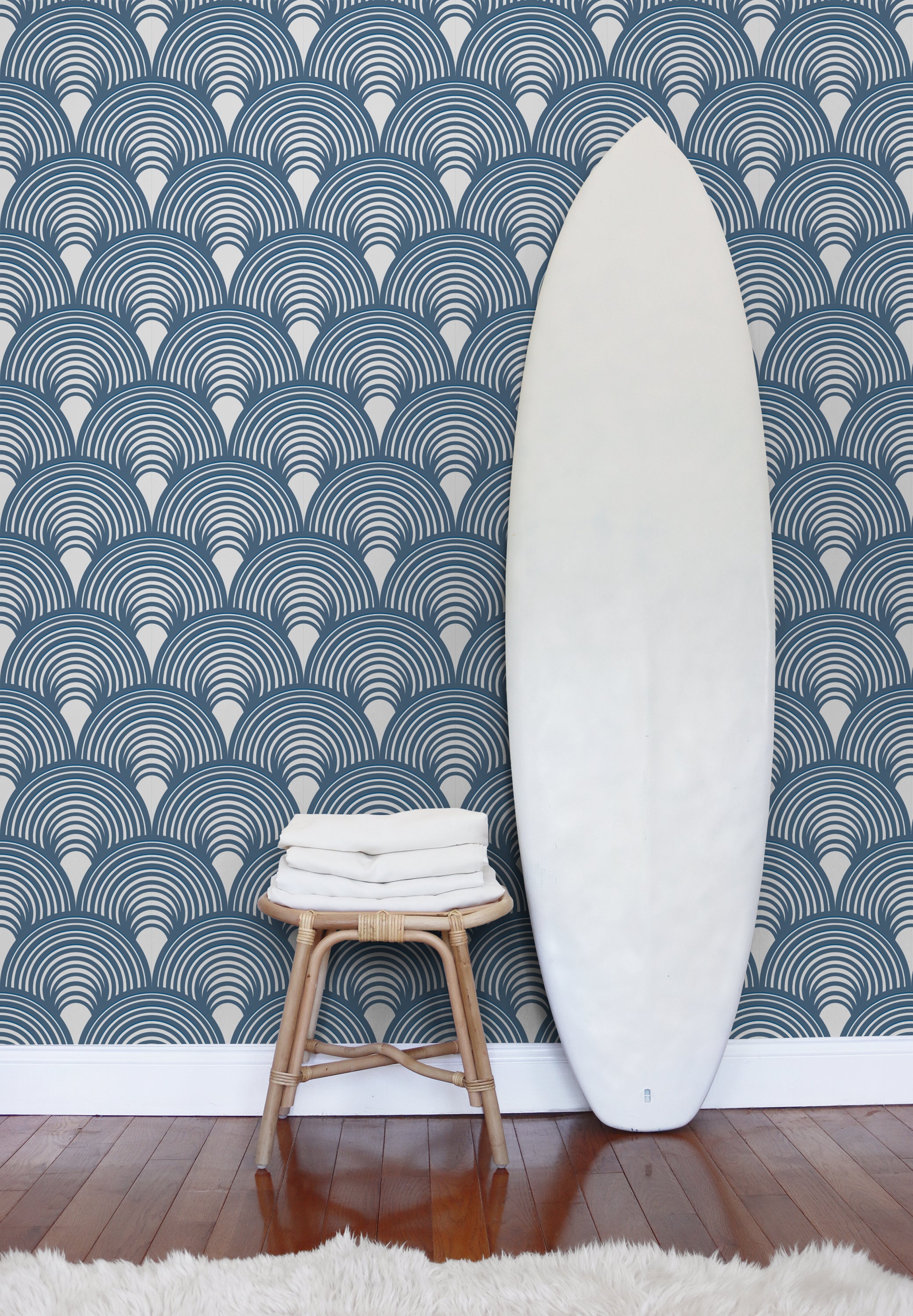 A tall white surfboard leans against a wall with blue and white scallop-pattern wallpaper designed Noemi Marozsan