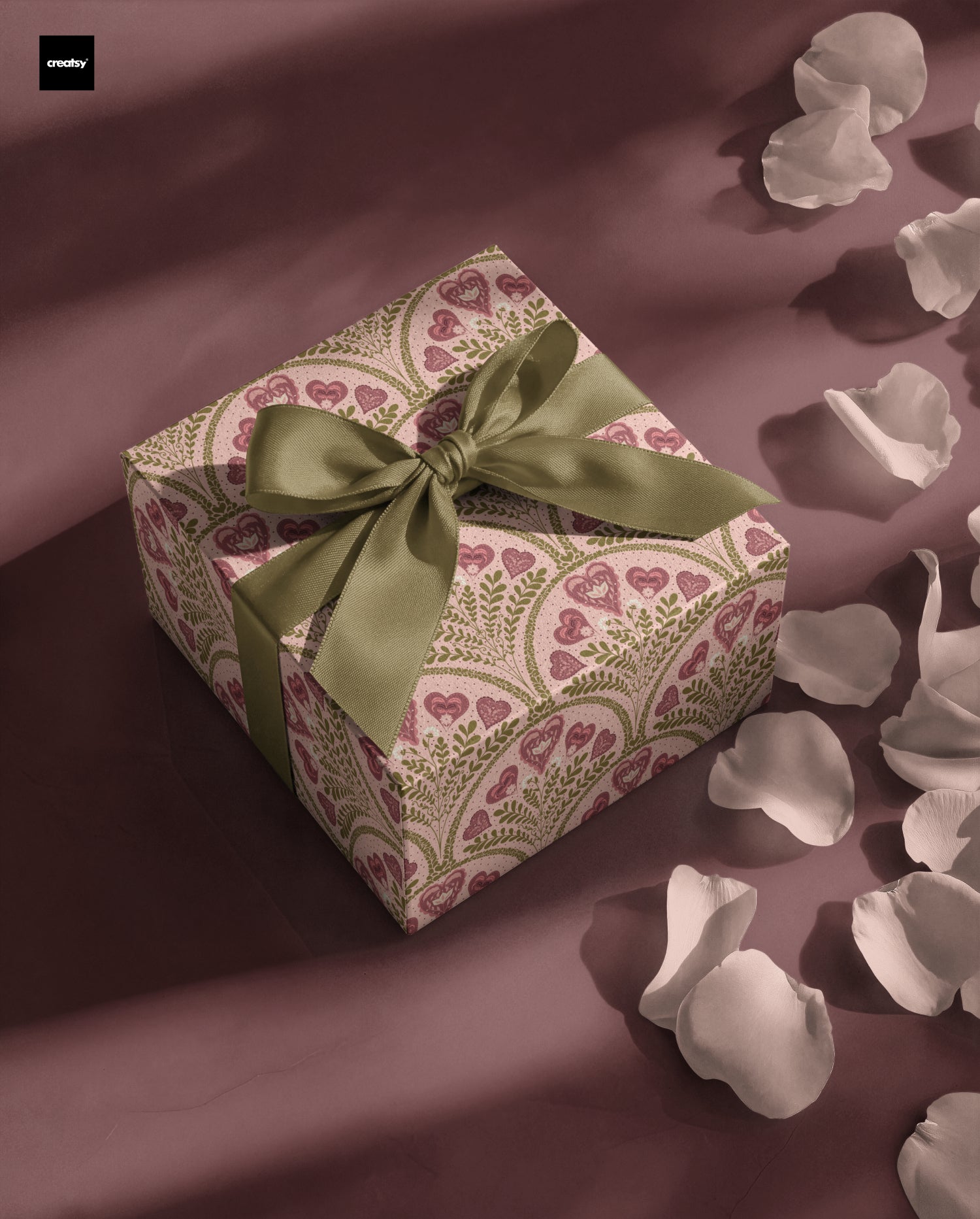 A square gift box wrapped in cream paper with a pink heart and paisley pattern, tied with an olive green satin ribbon, set on a dark mauve Scandinavian love design by Noemi Marozsan with scattered white flower petals.