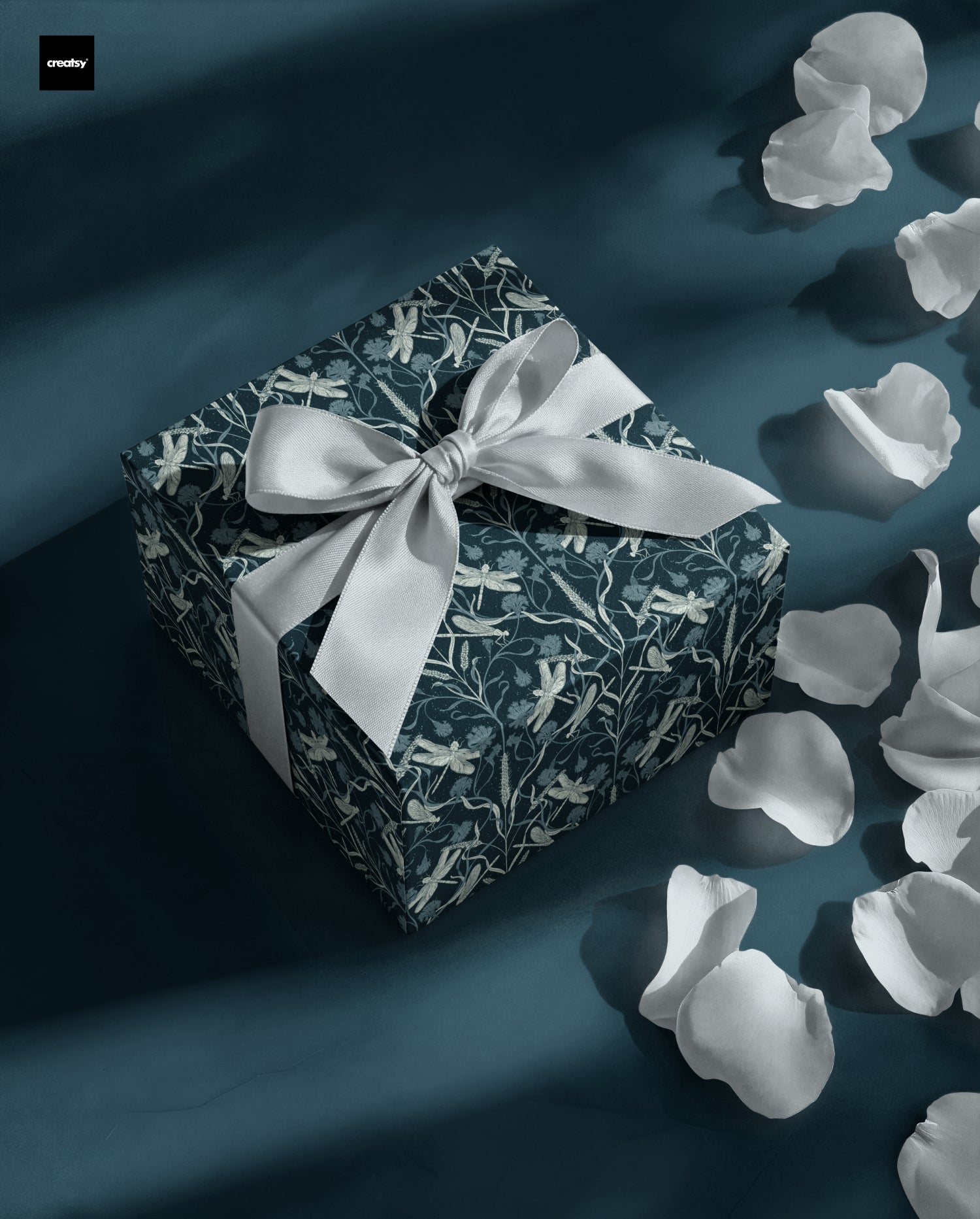 A square gift box wrapped in dark teal and white floral-patterned paper, tied with a large white satin bow, set on a dark teal dragonfly surface with scattered white flower petals. design by noemi marozsan