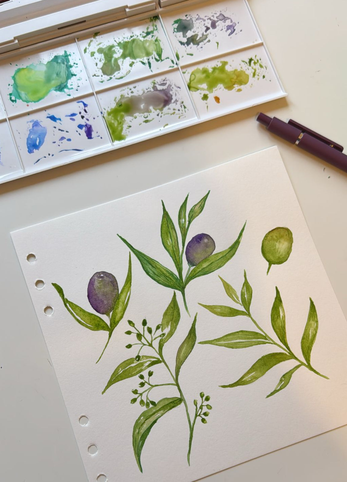 Watercolour painting in progress showing hand‑painted olive branches with green leaves and purple olives, surrounded by a paint palette and brush on an artist’s workspace, created by Noemi Marozsan Designs.