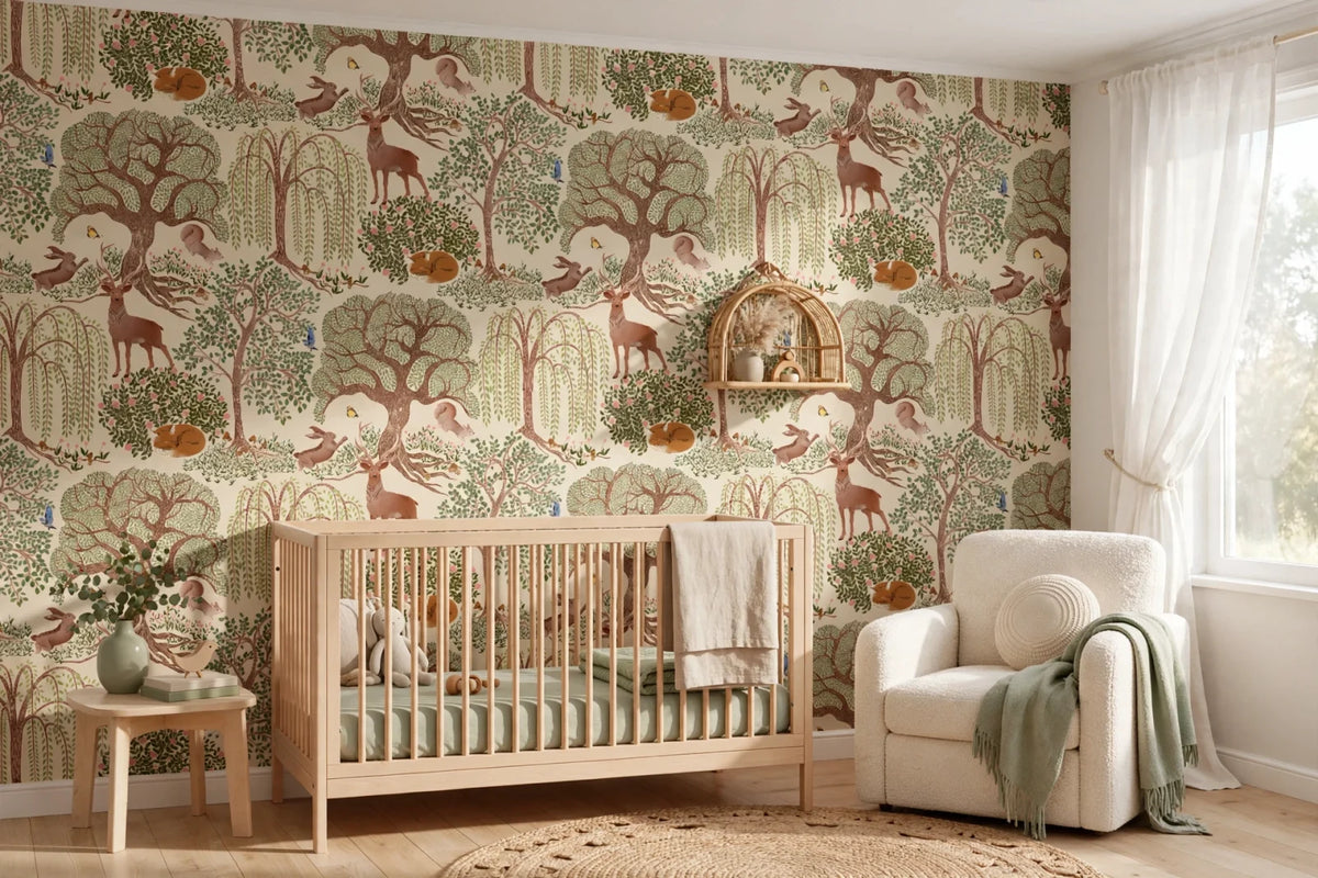 A styled nursery room featuring a wall covered in whimsical forest-themed wallpaper with deer, trees, and birds. In front of the wallpaper is a light wood crib, next to a cream armchair with a green throw blanket, a small wooden side table, and a window with sheer white curtains.